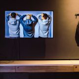 interior of art gallery with blak walls, showing tv screen with 3 men with hands on their heads. Next to it a transparent rucksack is hung up containing prosthetic hands