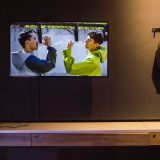 Interior of art gallery with black walls and tv showing 3 men with hands on their heads and transparent rucksack holding prosthetic hands
